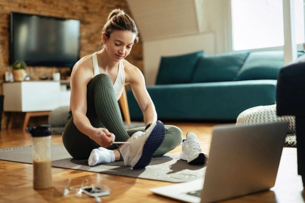 A woman getting ready to exercise at home, sitting in her living room on a yoga mat, putting on running shoes and using a laptop.